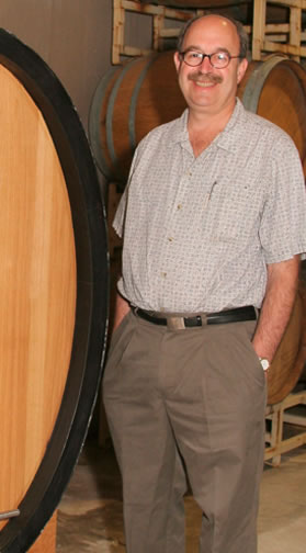 Man in a winery surrounded by large wooden wine barrels, smiling at the camera.